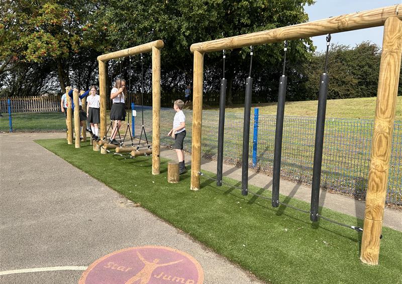 5 children are in a single file line to take on an obstacle course made from wood and rope. One child is on a step bridge as the other children are hopping across log stumps.
